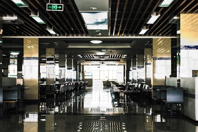 A spacious, modern cafeteria with rows of tables and chairs neatly arranged in a symmetrical pattern. The ceiling features a grid of rectangular lights and wooden beams, and there are reflective tiles on the floor adding to the symmetrical aesthetic. Natural light floods in from large windows at the end of the room, creating a bright and open atmosphere.