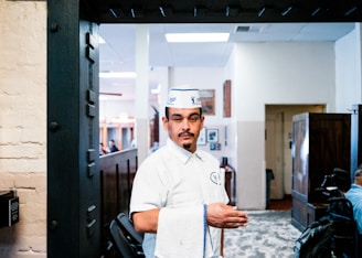 A person dressed in a white uniform and cap stands indoors, holding a towel over their arm. The background shows a dining area with wooden booths and a few people seated. The walls are decorated with framed photos and there is a mix of light and dark wood furnishings.
