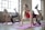 Three women are engaged in a fitness routine, following a workout in a bright, spacious studio with large windows. Each woman is on a pink exercise mat, performing a leg lift exercise, focusing on balance and strength.