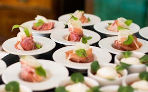 A collection of small white plates, each containing prosciutto wrapped around slices of cheese and garnished with fresh green leaves. The arrangement is neat and elegant, suitable for a catered event or gourmet tasting.