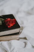 A single red rose is elegantly placed on top of two closed, vintage-looking books with black covers. The books rest on a soft, white fabric background.
