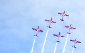 A formation of six red and white airplanes flying in an ascending diagonal line, leaving trails of white smoke against a clear blue sky.