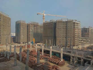 A large construction site with multiple high-rise buildings in various stages of completion. A crane is prominently positioned among the structures, and scaffolding surrounds the buildings. The foreground is filled with construction materials, including metal rods and wooden planks. The sky is clear and sunny.