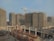 A large construction site with multiple high-rise buildings in various stages of completion. A crane is prominently positioned among the structures, and scaffolding surrounds the buildings. The foreground is filled with construction materials, including metal rods and wooden planks. The sky is clear and sunny.