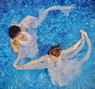 Two women are gracefully floating in a clear blue swimming pool, draped in delicate, semi-transparent fabric. The water creates a rippling effect around them, enhancing the ethereal and artistic composition.