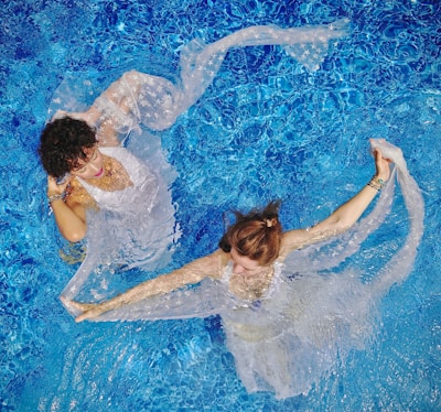 Two women are gracefully floating in a clear blue swimming pool, draped in delicate, semi-transparent fabric. The water creates a rippling effect around them, enhancing the ethereal and artistic composition.