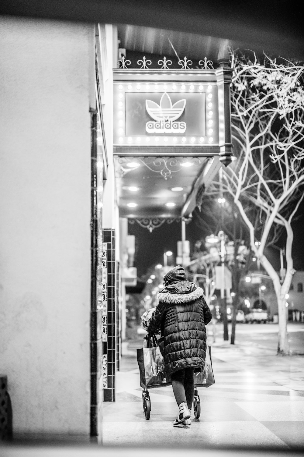 foto in scala di grigi di una persona che cammina sul marciapiede vicino al negozio Adidas