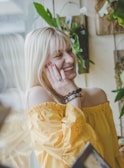 A woman is laughing with her hand gently touching her cheek. She is wearing an off-the-shoulder yellow top and has blonde hair. In the background, there are green plants hanging on the wall.