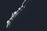 Bright neon light in the shape of a handwritten script, set against a dark background. The glowing light creates a striking contrast with the deep shadows, enhancing its luminescence.