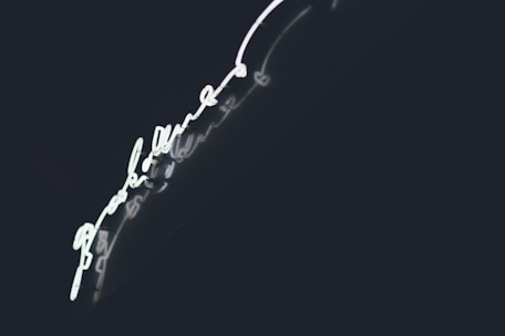 Bright neon light in the shape of a handwritten script, set against a dark background. The glowing light creates a striking contrast with the deep shadows, enhancing its luminescence.