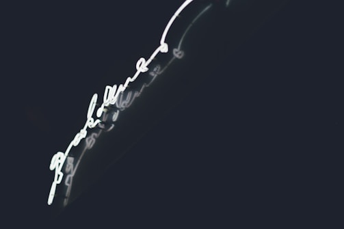 Bright neon light in the shape of a handwritten script, set against a dark background. The glowing light creates a striking contrast with the deep shadows, enhancing its luminescence.