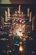 A long dining table is elegantly set with multiple wine glasses, silverware, and napkins standing upright. The lighting is dim and warm, creating a sophisticated and intimate atmosphere.