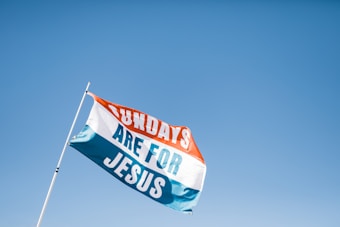 A flag with the words 'Sundays Are For Jesus' printed in large, bold letters. The flag has three horizontal stripes: orange, white, and blue. The flagpole is angled towards the left and is set against a clear blue sky, indicating a bright, sunny day.