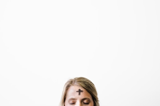 A person with their eyes closed has an ash cross on their forehead, symbolizing the observance of Ash Wednesday. The background is plain and light, emphasizing the solemn expression.
