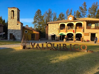 A rustic village scene with stone buildings, a clock tower, and a sign reading 'Valquirico' on a grassy area. The architecture combines stone and wooden elements, and several flags hang from a balcony. People are walking and enjoying the surroundings under a blue sky.