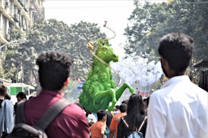 A large, vibrant green sculpture of a horse with multiple human faces along its body stands prominently in a busy outdoor setting. The sculpture is adorned with a decorative spiral element. People gather around, some with backpacks, observing the display. The background features tall trees and a few buildings, suggesting an urban setting.