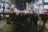 A group of people are marching down a city street at night, holding various protest signs and a rainbow flag. The street is lined with tall buildings, some of which are illuminated with lights. A notable sign reads 'CHICAGO,' prominently displayed in the background.