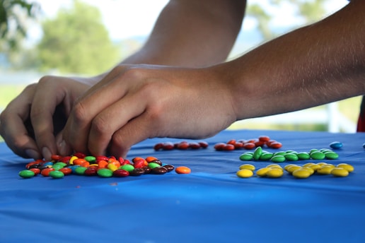Two hands are sorting colorful candy-coated chocolates on a blue tablecloth. The candies are arranged in small piles by color, including red, yellow, green, brown, and blue. The background is slightly blurred, indicating an outdoor setting with greenery.