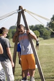 A group of people are outdoors on a sunny day, engaging in an activity involving wooden structures and ropes. One person is standing between two wooden poles with multiple ropes extending from the top, suggesting a team-building exercise or game.