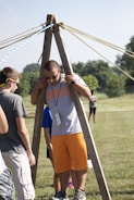 A group of people are outdoors on a sunny day, engaging in an activity involving wooden structures and ropes. One person is standing between two wooden poles with multiple ropes extending from the top, suggesting a team-building exercise or game.