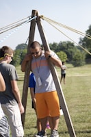A group of people are outdoors on a sunny day, engaging in an activity involving wooden structures and ropes. One person is standing between two wooden poles with multiple ropes extending from the top, suggesting a team-building exercise or game.