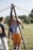 A group of people are outdoors on a sunny day, engaging in an activity involving wooden structures and ropes. One person is standing between two wooden poles with multiple ropes extending from the top, suggesting a team-building exercise or game.