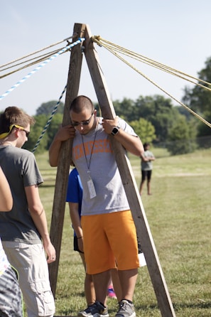 A group of people are outdoors on a sunny day, engaging in an activity involving wooden structures and ropes. One person is standing between two wooden poles with multiple ropes extending from the top, suggesting a team-building exercise or game.