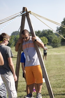 A group of people are outdoors on a sunny day, engaging in an activity involving wooden structures and ropes. One person is standing between two wooden poles with multiple ropes extending from the top, suggesting a team-building exercise or game.