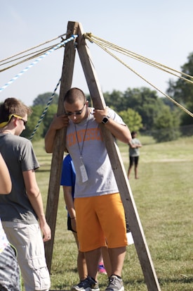 A group of people are outdoors on a sunny day, engaging in an activity involving wooden structures and ropes. One person is standing between two wooden poles with multiple ropes extending from the top, suggesting a team-building exercise or game.