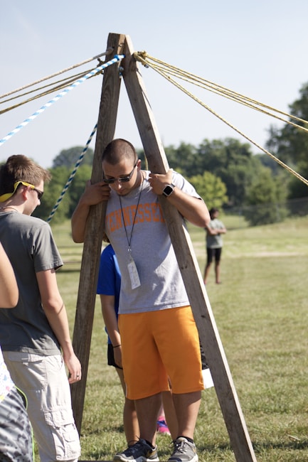 A group of people are outdoors on a sunny day, engaging in an activity involving wooden structures and ropes. One person is standing between two wooden poles with multiple ropes extending from the top, suggesting a team-building exercise or game.