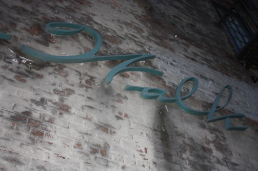 A brick wall with slightly faded paint displaying an artistic script sign in greenish-blue letterings. The surface appears weathered, with areas of discoloration and texture variation adding an urban aesthetic.