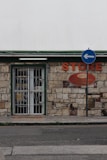 A small store with a stone facade features a green-trimmed door and metal security gate. An orange sign above reads 'STORE' beside a directional street sign with a blue background and white arrow. The building has an aged, rustic look with varying shades of stone and some brickwork.