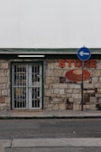 A small store with a stone facade features a green-trimmed door and metal security gate. An orange sign above reads 'STORE' beside a directional street sign with a blue background and white arrow. The building has an aged, rustic look with varying shades of stone and some brickwork.