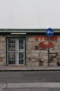 A small store with a stone facade features a green-trimmed door and metal security gate. An orange sign above reads 'STORE' beside a directional street sign with a blue background and white arrow. The building has an aged, rustic look with varying shades of stone and some brickwork.