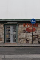A small store with a stone facade features a green-trimmed door and metal security gate. An orange sign above reads 'STORE' beside a directional street sign with a blue background and white arrow. The building has an aged, rustic look with varying shades of stone and some brickwork.