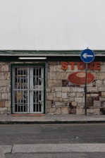 A small store with a stone facade features a green-trimmed door and metal security gate. An orange sign above reads 'STORE' beside a directional street sign with a blue background and white arrow. The building has an aged, rustic look with varying shades of stone and some brickwork.