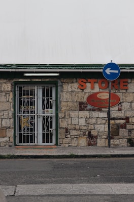 A small store with a stone facade features a green-trimmed door and metal security gate. An orange sign above reads 'STORE' beside a directional street sign with a blue background and white arrow. The building has an aged, rustic look with varying shades of stone and some brickwork.