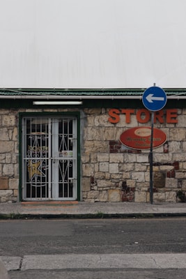 A small store with a stone facade features a green-trimmed door and metal security gate. An orange sign above reads 'STORE' beside a directional street sign with a blue background and white arrow. The building has an aged, rustic look with varying shades of stone and some brickwork.