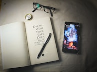 An open book lies on a surface next to a smartphone, a pair of eyeglasses, a pen, and a small compass. The book's title page reads 'Dream With Your Eyes Open' by Ronnie Screwvala. The smartphone displays a lock screen with the word 'Coffee' in a stylized smoke design.