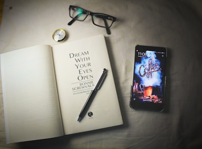 An open book lies on a surface next to a smartphone, a pair of eyeglasses, a pen, and a small compass. The book's title page reads 'Dream With Your Eyes Open' by Ronnie Screwvala. The smartphone displays a lock screen with the word 'Coffee' in a stylized smoke design.