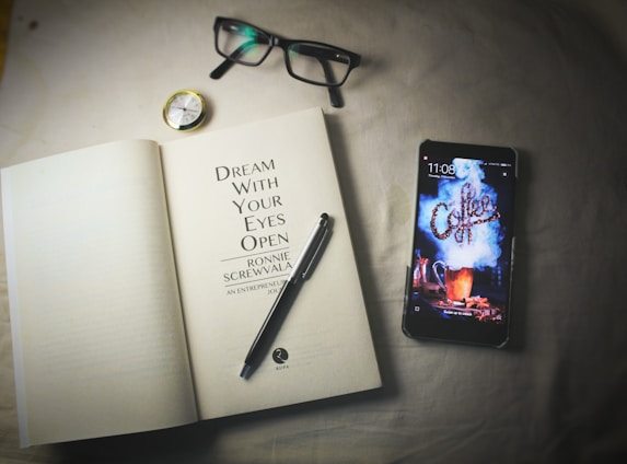 An open book lies on a surface next to a smartphone, a pair of eyeglasses, a pen, and a small compass. The book's title page reads 'Dream With Your Eyes Open' by Ronnie Screwvala. The smartphone displays a lock screen with the word 'Coffee' in a stylized smoke design.