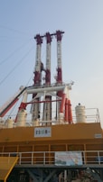 A large industrial structure with several tall red and white vertical components extending into the sky. The base is a solid yellow with various railings and signage. There is also a netting system and several cylindrical structures, indicating a complex setup likely used for construction or industrial purposes.