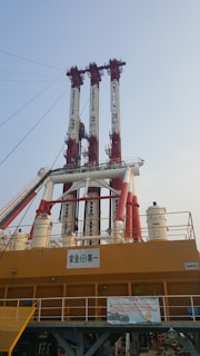 A large industrial structure with several tall red and white vertical components extending into the sky. The base is a solid yellow with various railings and signage. There is also a netting system and several cylindrical structures, indicating a complex setup likely used for construction or industrial purposes.