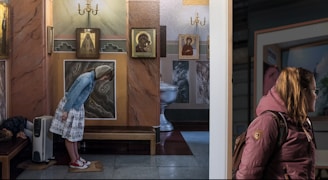 A woman in a denim jacket and skirt bows in front of religious icons on a wall. A child sleeps on a bench beside her. There is a heater nearby. Another woman wearing a jacket with a backpack stands in the foreground, looking back towards the scene.