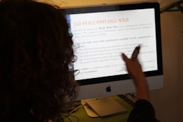 A person with curly hair is pointing at a computer screen displaying an Italian text about the development of the web. The person is holding a pen, indicating they may be discussing or presenting the content.