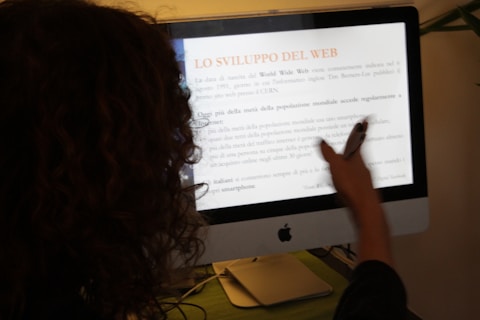 A person with curly hair is pointing at a computer screen displaying an Italian text about the development of the web. The person is holding a pen, indicating they may be discussing or presenting the content.