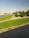 A well-maintained park area featuring carefully trimmed circular bushes and expansive green lawns adjacent to a road. The scene includes a clear sky and trees in the background, with a few vehicles visible on the road.