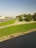 A well-maintained park area featuring carefully trimmed circular bushes and expansive green lawns adjacent to a road. The scene includes a clear sky and trees in the background, with a few vehicles visible on the road.