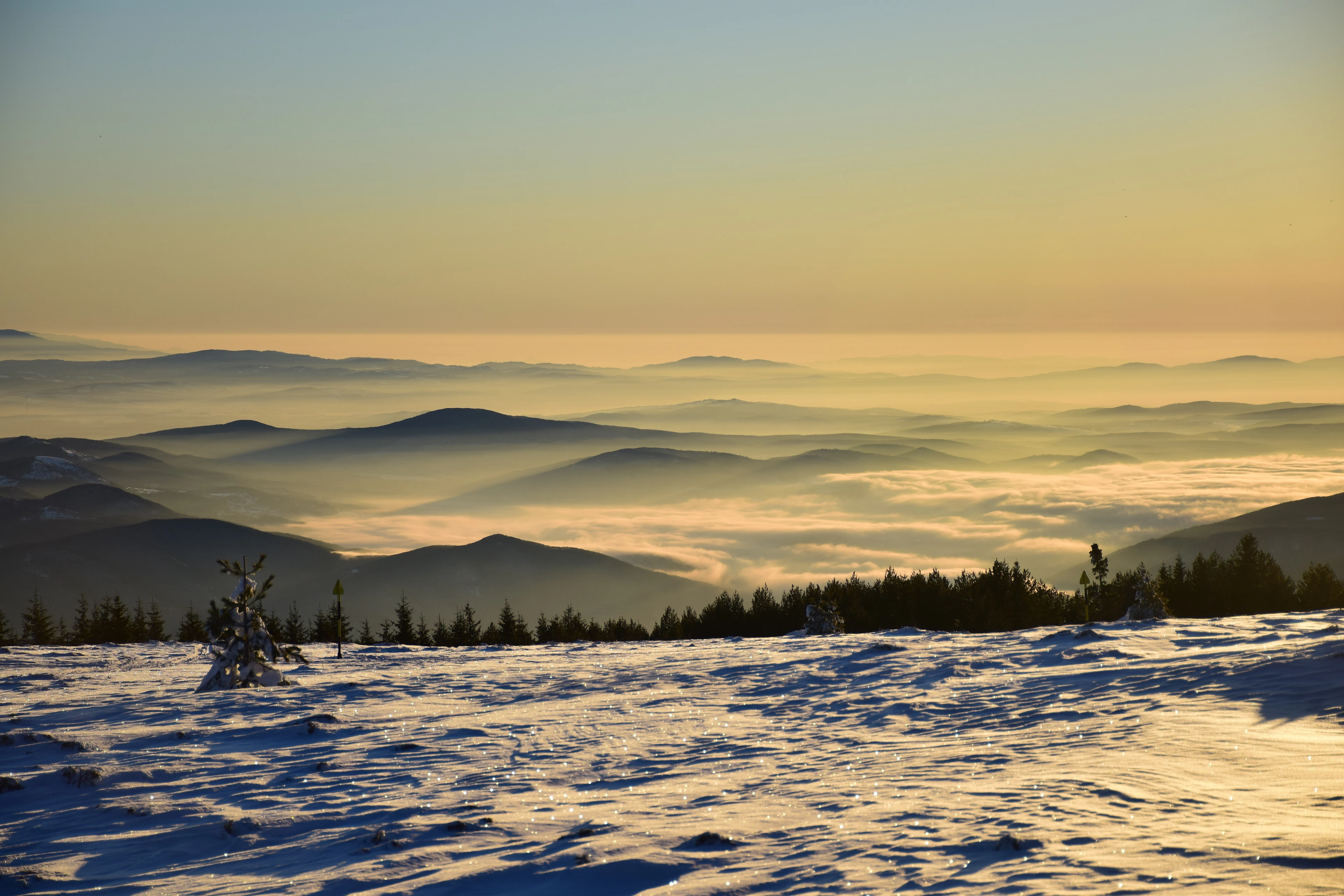 Sunrise from the snowfields of Vitosha above Sofia, with a sea of cloud filling the valleys and layered blue ridges receding into a gold horizon.