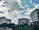 Several modern office buildings are positioned against a backdrop of a partly cloudy sky. The architectural structures are surrounded by greenery, including a well-maintained lawn and various plants, adding to the urban landscape.